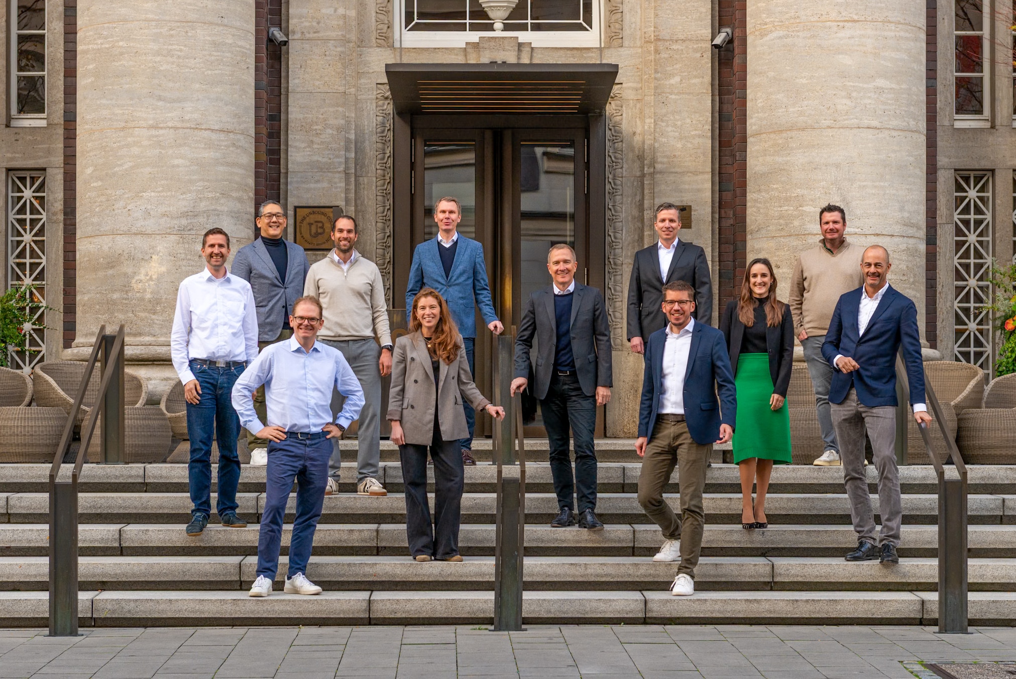 A team of twelve colleagues posed on the steps of an ornate historic building, standing in a relaxed formation and smiling at the camera.